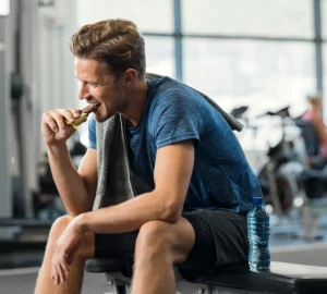 a man eating a protein bar while sitting on a bench in the gym