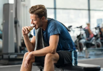 a man eating a protein bar while sitting on a bench in the gym