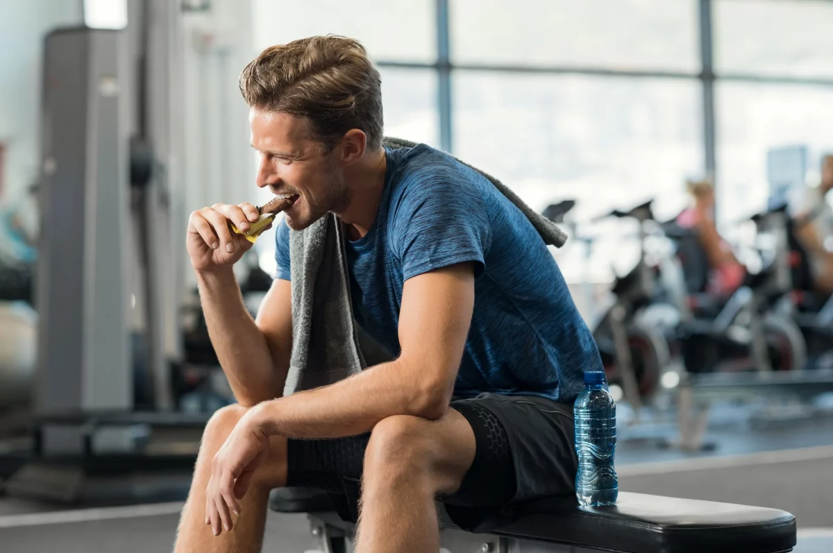 a man eating a protein bar while sitting on a bench in the gym