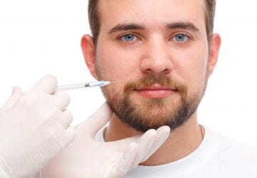 A male doctor is injected by a doctor wearing white gloves, botox on the cheek near the mouth. Photo on white background