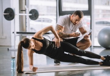 Male personal trainer looking at timer and young athletic woman doing side plank on fitness mat