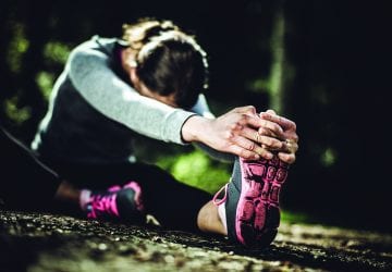 A dark moody image of a woman in her early 30's stretching her legs in a bright Pacific Northwest forest after a jog. Selective focus on her shoe and hands in the foreground, her shoes dirty from a gruelling run.