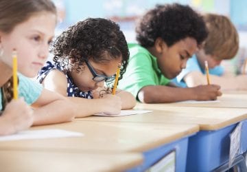 Multi-ethnic elementary school children writing in classroom. Focus on Hispanic girl wearing eyeglasses (8-9 years).