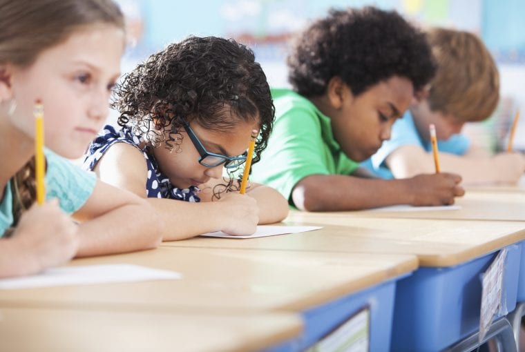 Multi-ethnic elementary school children writing in classroom. Focus on Hispanic girl wearing eyeglasses (8-9 years).
