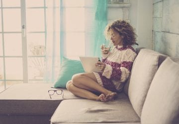 beautiful middle age woman with curly hair working at home and relaxing on the sofa with a tablet and internet. drinking tea or lemonade. happy relaxed leisure activity indoor