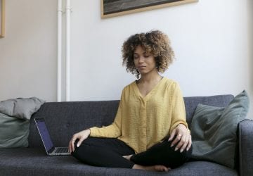 Young woman sitting on couch at home next to laptop meditating
