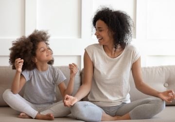 Excited woman teaching cute little adorable daughter meditating and practicing yoga exercises. Happy mixed race family sitting on comfy couch in lotus position, having fun together.
