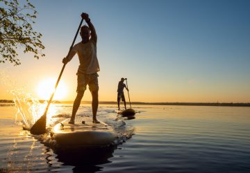 Men, friends sail on a SUP boards in a rays of rising sun. Stand up paddle boarding - awesome active recreation in nature. Backlight.
