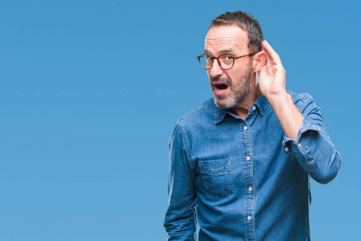 a man against a blue background cupping his hand to his ear to be able to hear better