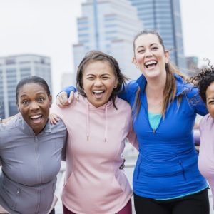 A multi-ethnic group of five women standing together on a city waterfront, smiling at the camera. They are in their 30s and 40s.
