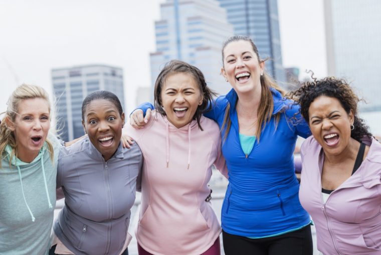 A multi-ethnic group of five women standing together on a city waterfront, smiling at the camera. They are in their 30s and 40s.