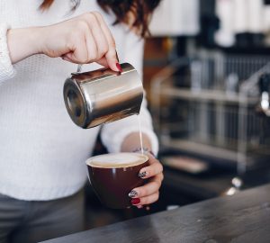 A girl pours milk foam into coffee. Waitress prepares coffee. Woman is pouring milk into coffee cup