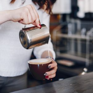A girl pours milk foam into coffee. Waitress prepares coffee. Woman is pouring milk into coffee cup