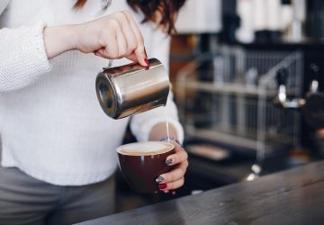 A girl pours milk foam into coffee. Waitress prepares coffee. Woman is pouring milk into coffee cup